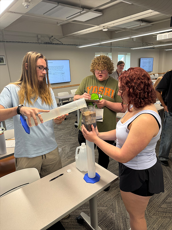 Students conduct a hands-on water filtration demonstration in a classroom, pouring water through testing equipment while classmates observe and take notes.