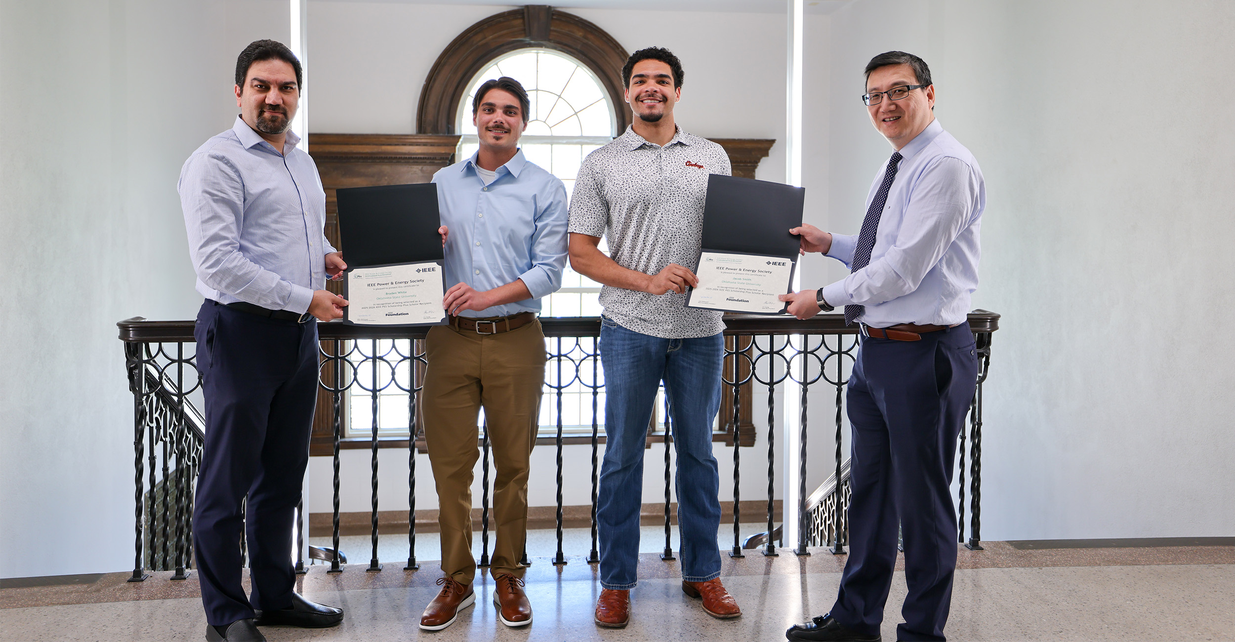 Four people stand side by side on an interior balcony holding open certificate folders toward the camera, posing for a formal award presentation with a large arched window and railing visible behind them.