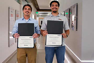 Two students stand side by side in a hallway holding framed certificates, posing for a recognition photo with academic posters and corridor walls visible behind them.