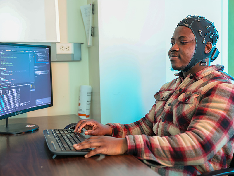 A man wearing an electrode cap sits infront of a computer.