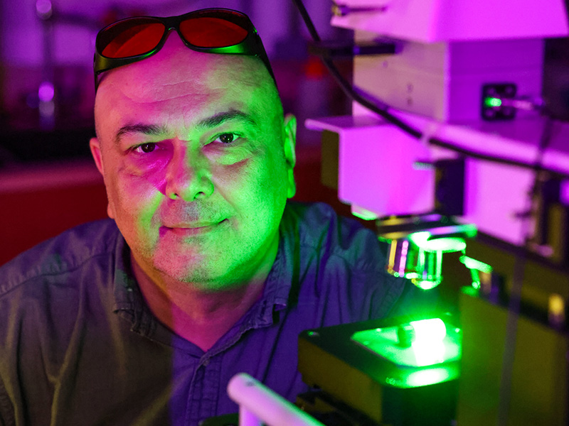 A researcher in a lab sits infront of a spectroscopy machine with laser googles resting on the top of his head. The background is illuminating with purple lights and the glow from the green laser can be seen across the researchers face.