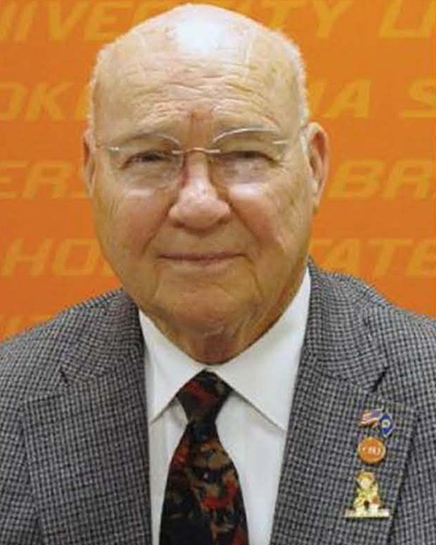 A man with glasses in a checkered suit jacket, white shirt, and patterned tie, standing in front of an orange background with partial text.
