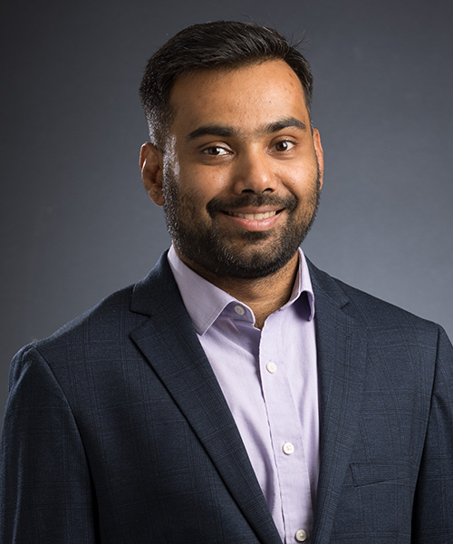 Professional portrait of a smiling man wearing a dark navy suit jacket and light purple button-down shirt, posed against a dark gray studio background.