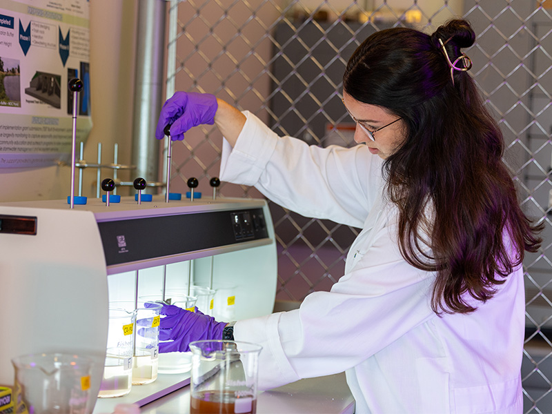 Person in a lab coat and gloves adjusts laboratory equipment while working with labeled beakers inside a bench-top testing unit.