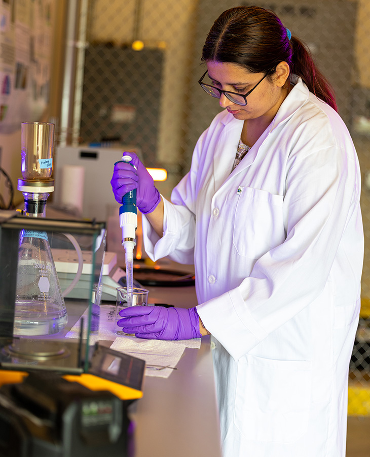 Person in a lab coat and gloves uses a pipette to transfer liquid into a beaker at a laboratory workbench.