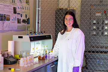 Person in a lab coat and gloves uses a pipette to transfer liquid into a beaker at a laboratory workbench.