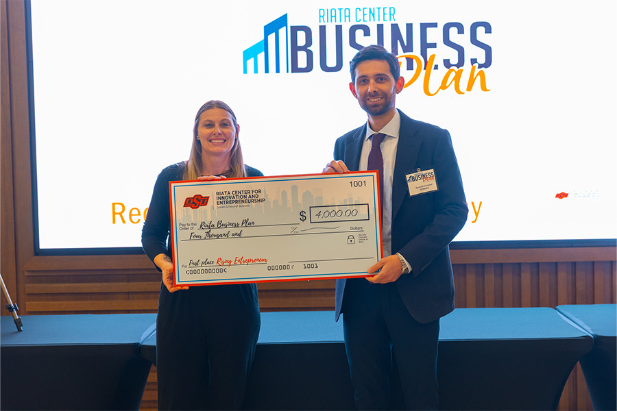 Two people smile as they pose with a giant check presented during the 2026 Riata Business Plan Competition. They are photographed wearing business casual clothing on a stage under a spotlight.