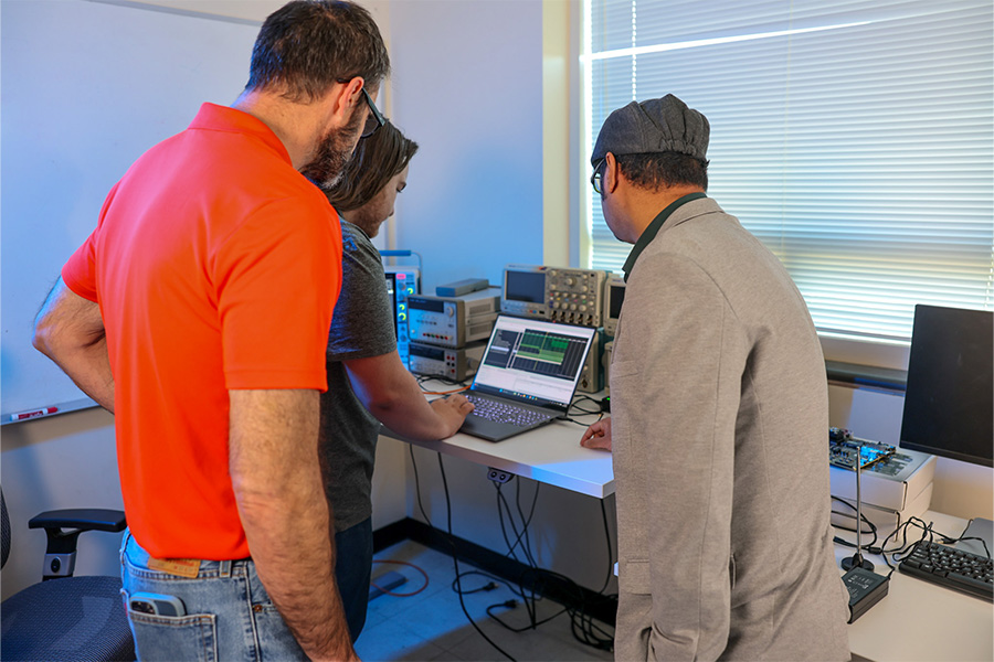 A laboratory scene shows three people gathered around a workstation with electronic testing equipment.