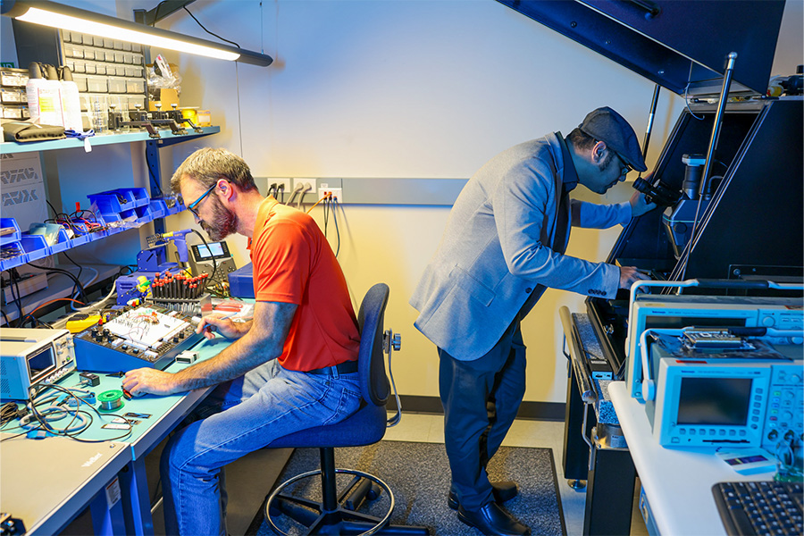 A brightly lit electronics laboratory with two people working at separate stations.