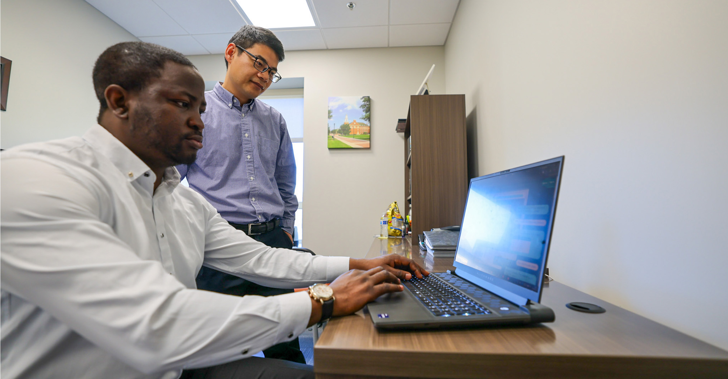 Two colleagues collaborate in an office, with one typing on a laptop displaying code while the other stands nearby observing, illustrating hands‑on database or programming work.