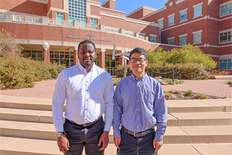 Two people stand side by side on steps outside an Oklahoma State University engineering building, with brick academic facilities and landscaped walkways visible in the background, illustrating a campus setting.