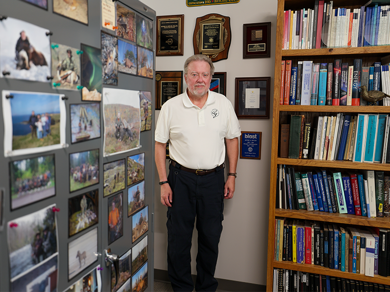 Portrait of a person standing in an office beside framed awards and certificates, with engineering textbooks visible on a bookshelf in the foreground.