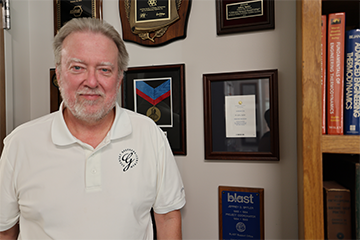 Person standing in an office between a display of field and research photographs and a bookshelf filled with engineering and science texts. Awards and certificates are mounted on the wall behind the person.