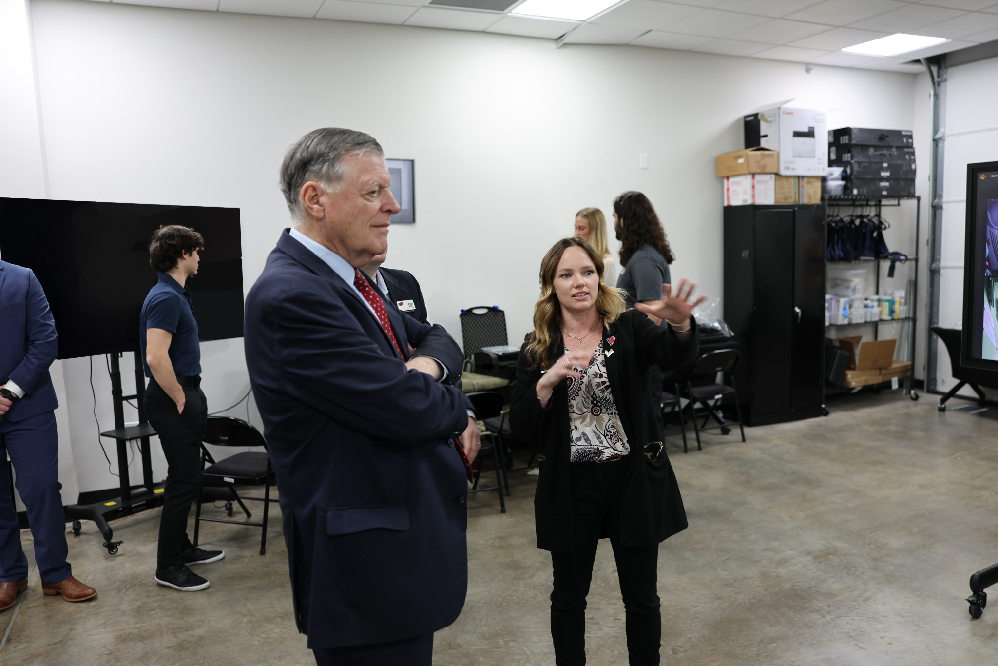 People stand in a technology lab or demonstration space as one person gestures while explaining equipment and displays, illustrating a guided tour or discussion of facilities and capabilities.