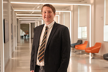Portrait of a person in business attire standing in a modern campus hallway with geometric light fixtures and lounge seating, highlighting a professional academic setting.