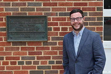 Dr. Tony McAleavy smiles while standing beside a historic bronze plaque marking the Fire Service Training School.