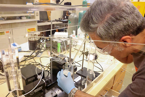 A researcher wearing safety glasses and blue nitrile gloves adjusts an experimental apparatus inside a clear protective enclosure in a laboratory