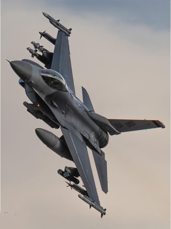 A gray military fighter jet banking sharply in midair, carrying multiple missiles under its wings against a pale sky.