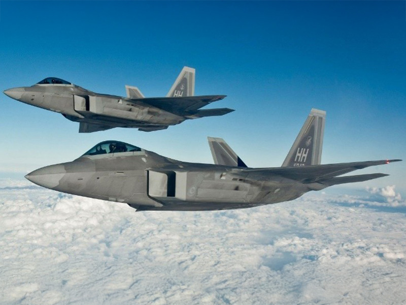 Two gray stealth fighter jets flying in formation above a blanket of white clouds under a blue sky.