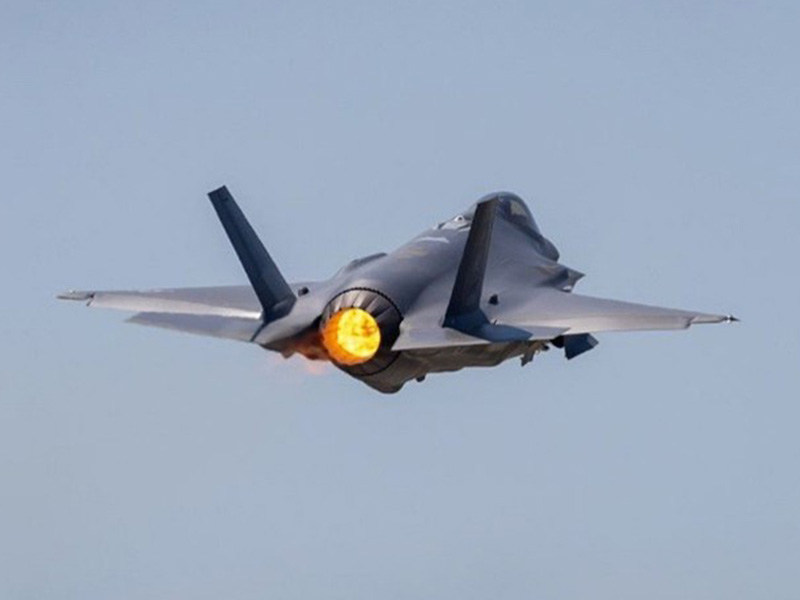 A modern stealth fighter jet viewed from behind with its engine afterburner glowing brightly as it flies against a clear sky.