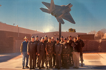 Group of people standing outdoors in front of a mounted fighter jet display on a pole, with a brick building visible in the background.