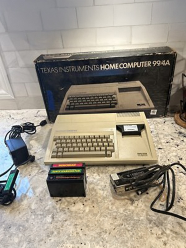 Vintage Texas Instruments TI-99/4A home computer displayed on a countertop with original box, power cords, and cartridge accessories arranged nearby.