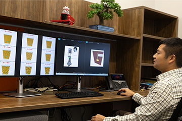 Researcher seated at a desk in an office with dual computer monitors displaying particle simulation models and engineering diagrams. Wood shelving, a potted tree, and office equipment are visible in the workspace.