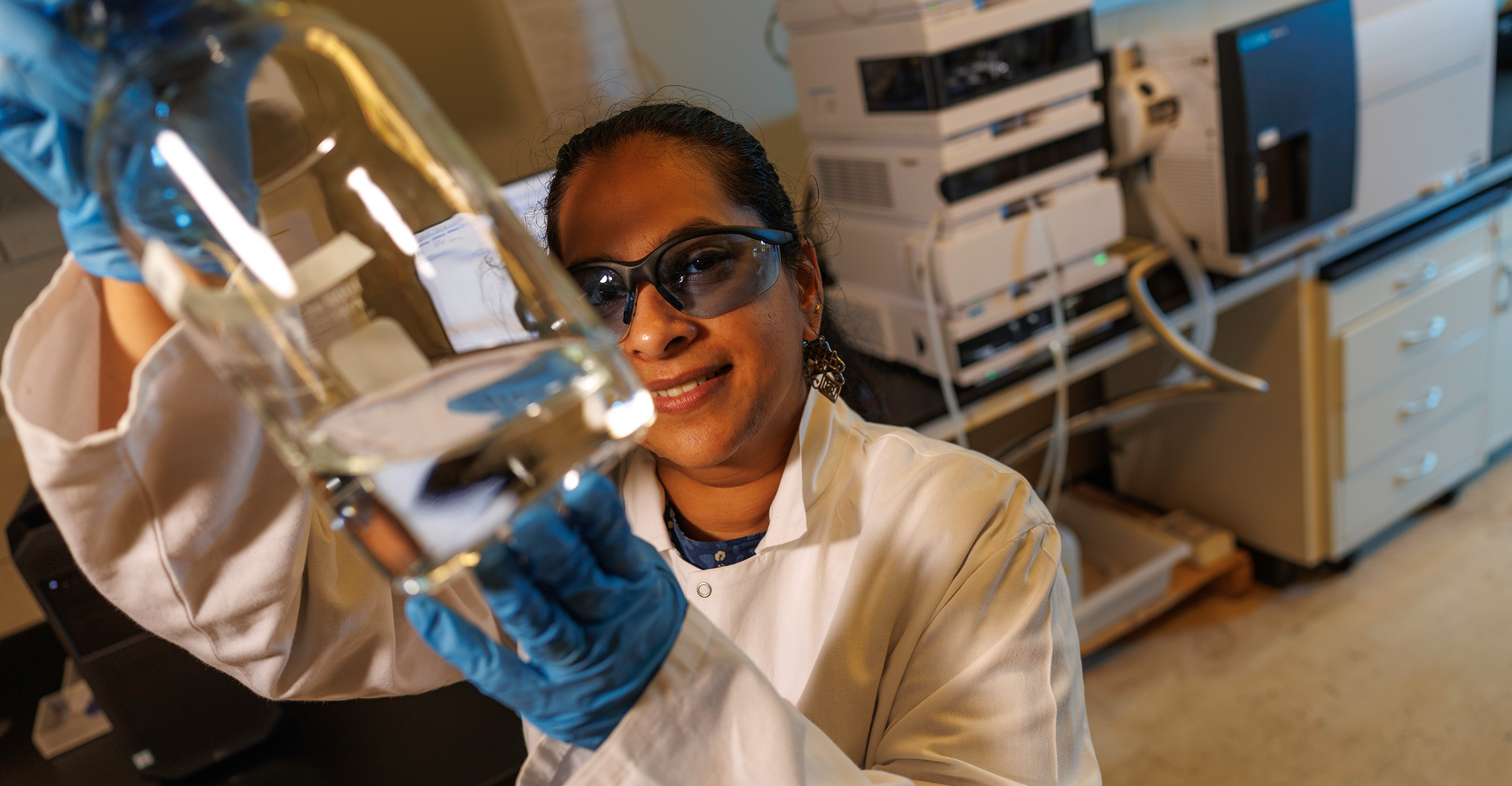 Researcher in a laboratory wearing safety glasses and gloves examines a glass container filled with liquid, with analytical instruments visible in the background.