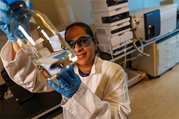Researcher in a laboratory wearing safety glasses and gloves examines a glass container filled with liquid, with analytical instruments visible in the background.