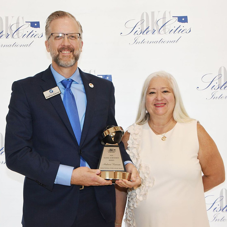 Two individuals pose with a Global Achievement Award at a Sister Cities International event, with one holding the trophy in front of a backdrop displaying the organization’s logo.