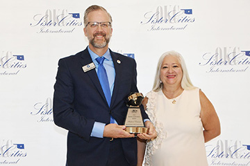 Two individuals pose with a Global Achievement Award at a Sister Cities International event, with one holding the trophy in front of a backdrop displaying the organization’s logo.