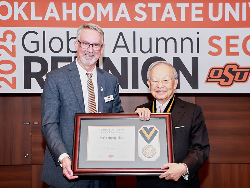 Two individuals are standing in front of a banner that reads 'OKLAHOMA STATE UNIV 2025 Global Alumni Reunion.' One person is holding a framed certificate with a medal inside. The Oklahoma State University logo is visible on the right side of the banner.