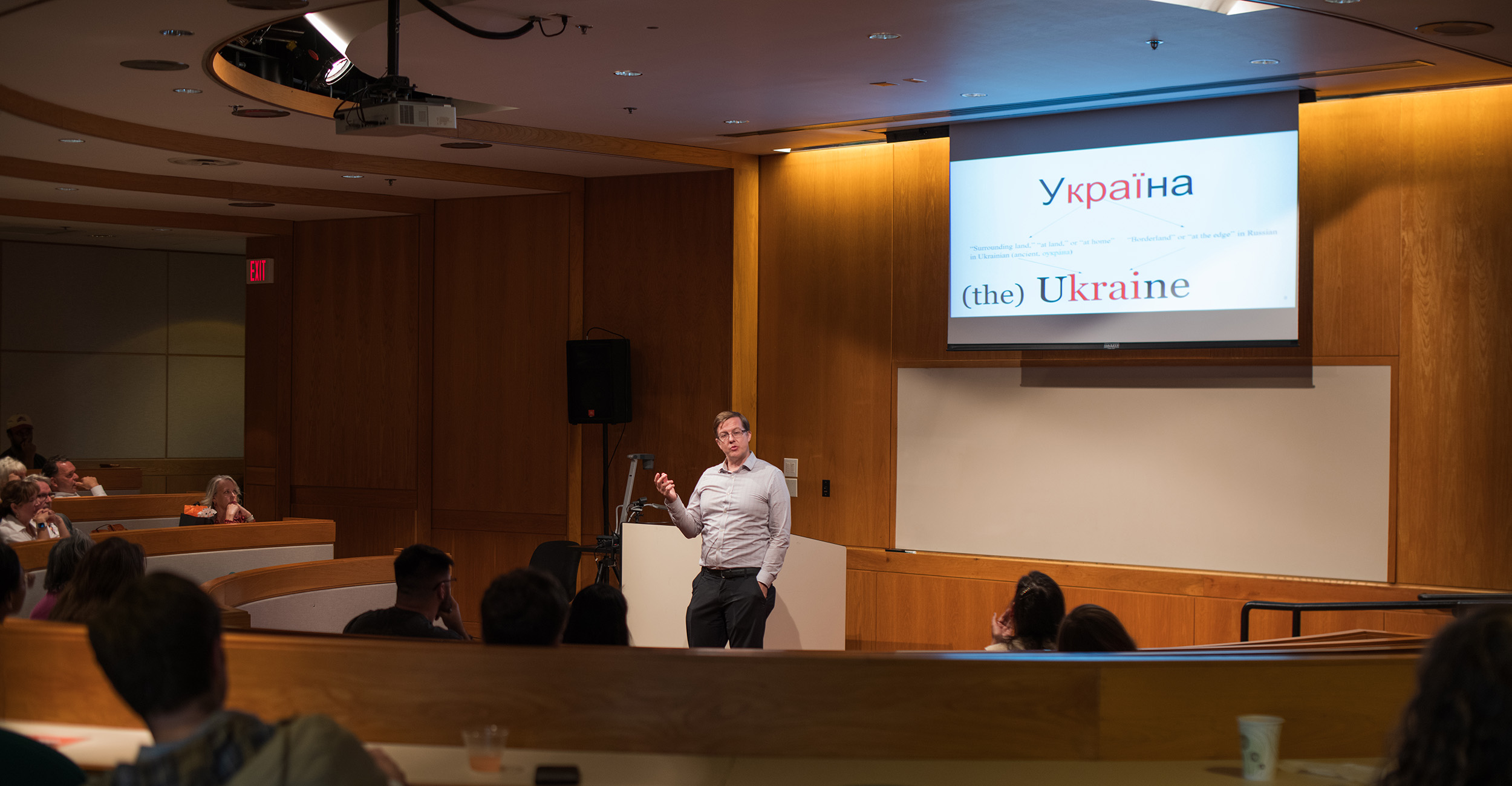 A presenter stands at a podium in a lecture hall, addressing an audience seated in curved rows. Behind the presenter, a large screen displays a slide with the word "Україна" (Ukraine) and explanatory text in English about the correct usage of "Ukraine" versus "the Ukraine.