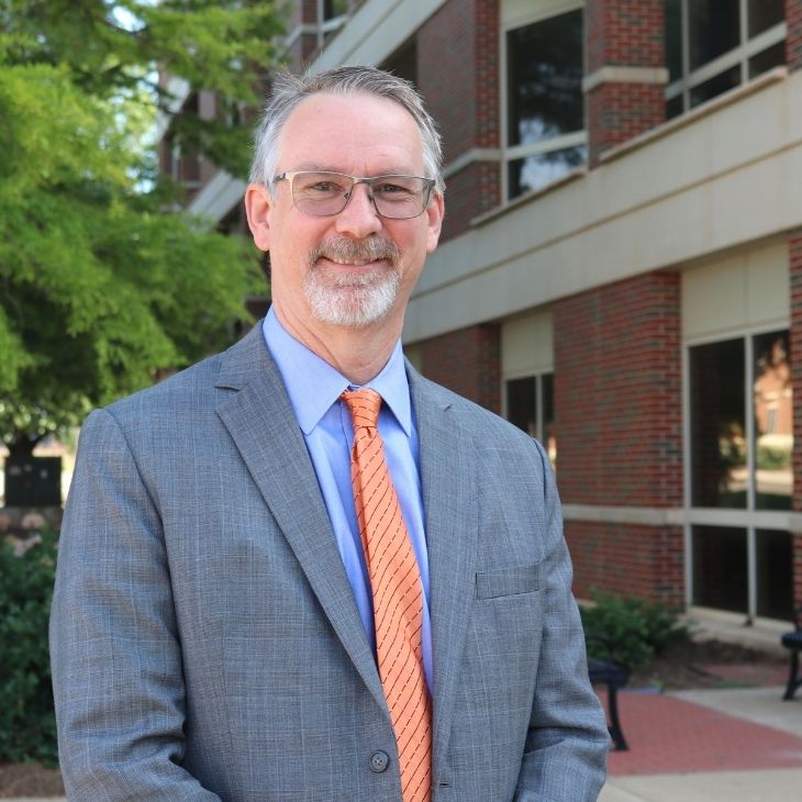 A Caucasian man stands with a suit on in front of a building.