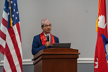 A speaker stands behind a podium holding a microphone, framed by the United States and Nepalese flags during a visit to Oklahoma State University.