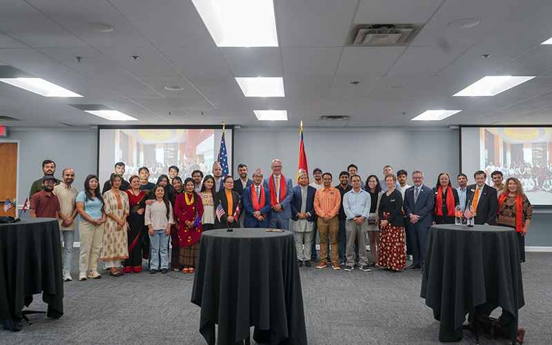 A large group gathers for a photo in a reception room with U.S. and Nepalese flags displayed behind them.