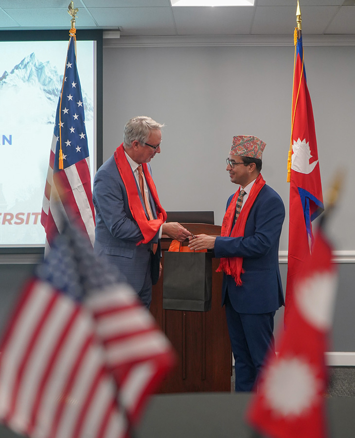 Two people exchange a gift bag at the front of a room between the United States and Nepalese flags.