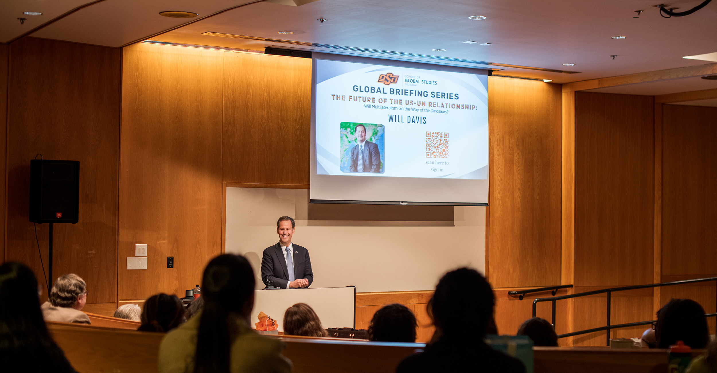 A speaker stands at a podium addressing an audience in a lecture hall during an OSU Global Briefing Series event, with a presentation slide about the future of the U.S.–UN relationship displayed behind him.