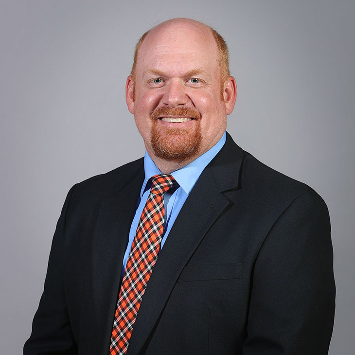 Professional head-and-shoulders portrait of a person wearing a dark suit jacket, light blue shirt and orange plaid tie against a plain gray background.