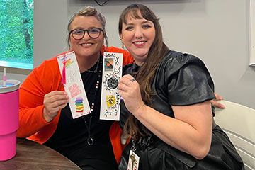 Two people sit side by side indoors, smiling at the camera while holding decorative bookmarks with book-themed designs, highlighting a reading or book club activity.