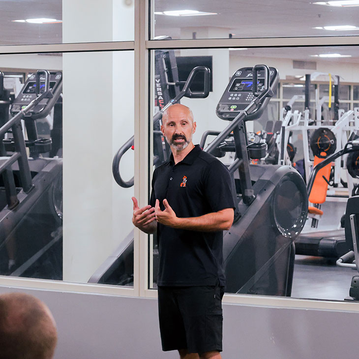 An instructor stands in a gym speaking to a group while exercise equipment such as treadmills and weight machines are visible behind.