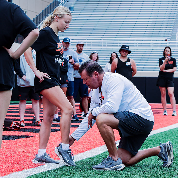 A coach kneels on a turf field adjusting an athlete’s ankle position while a group of participants observes during an outdoor training clinic in a stadium.