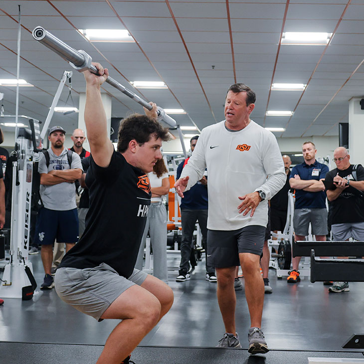 A coach demonstrates proper lifting form by guiding an athlete performing an overhead squat with a barbell while participants observe in a weight room.