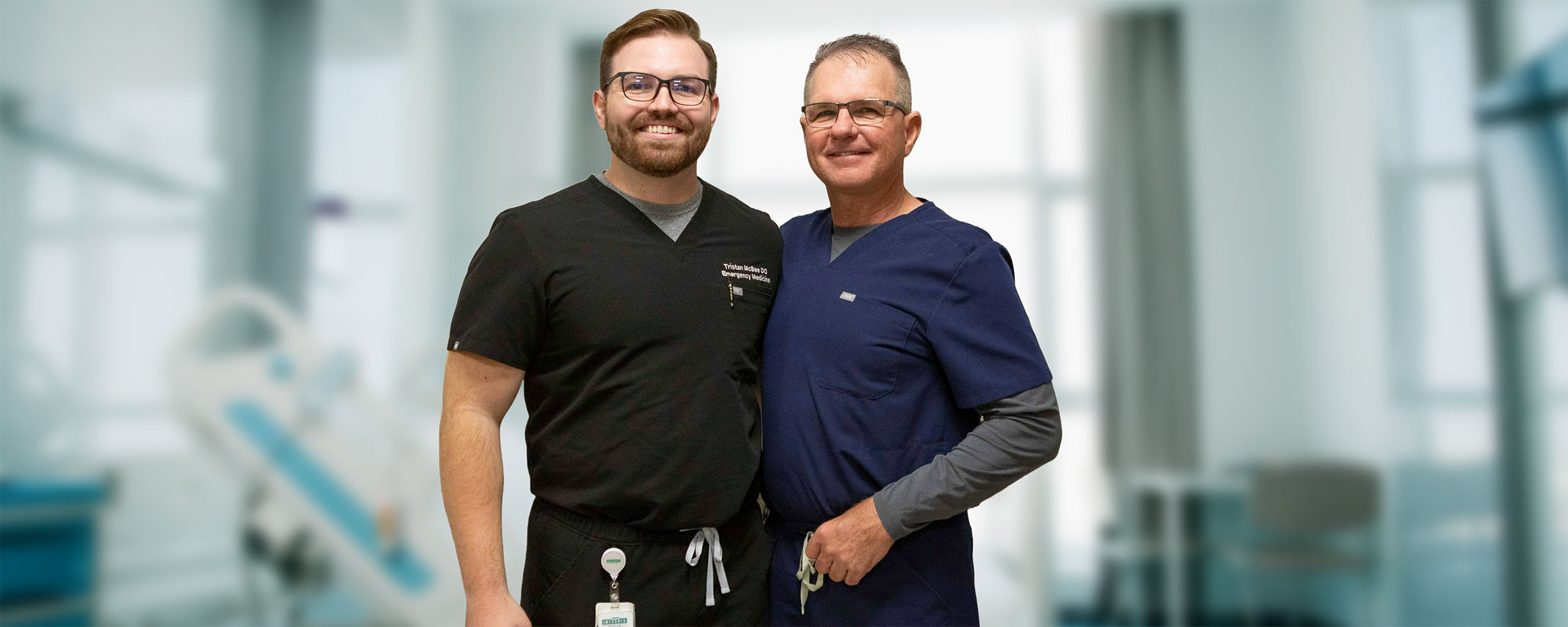 Two people wearing medical scrubs stand side by side in a bright clinical setting, with medical equipment visible in the softly blurred background.