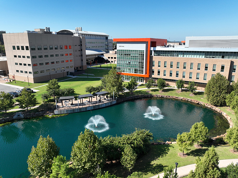 An aerial view of a modern campus with several large academic buildings surrounding a landscaped pond with three water fountains. Green lawns, trees, and walking paths fill the space between the buildings, set against a bright blue sky.