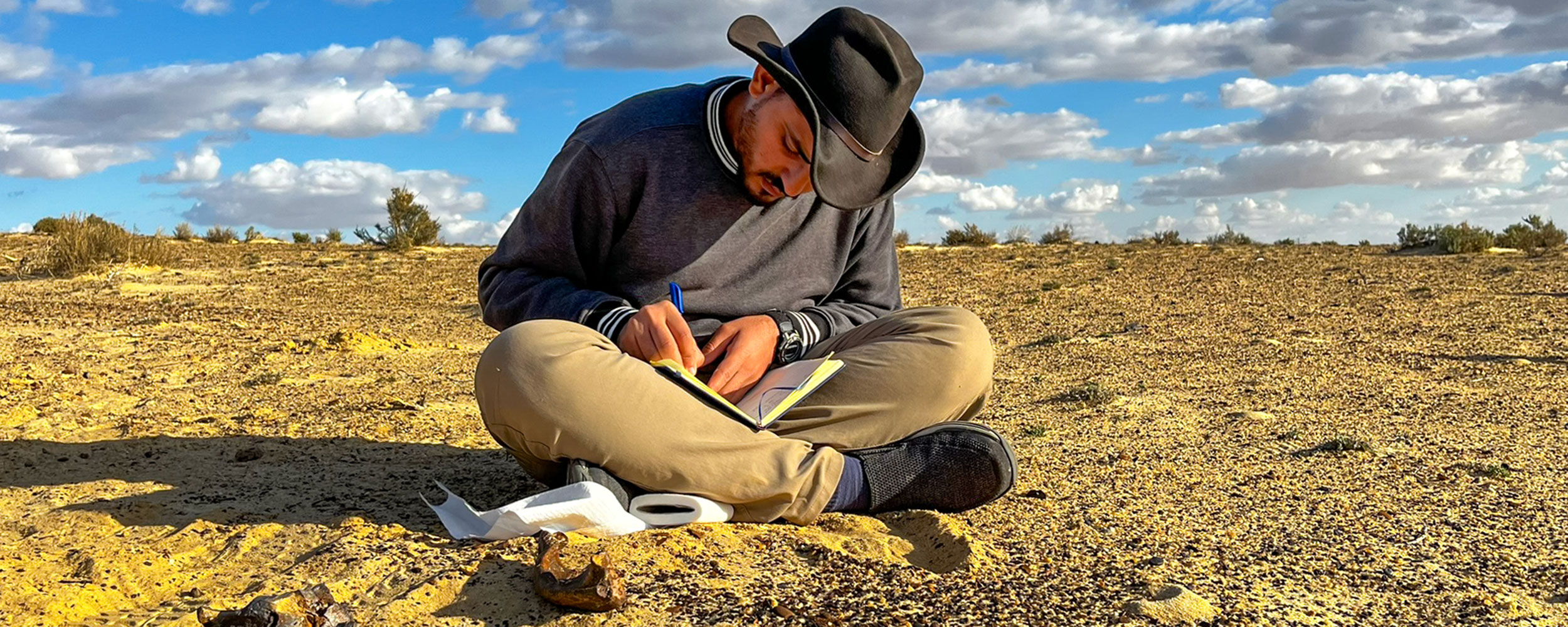 A person sits cross‑legged on a dry, open landscape, writing in a notebook, illustrating field research or scientific note‑taking conducted outdoors.
