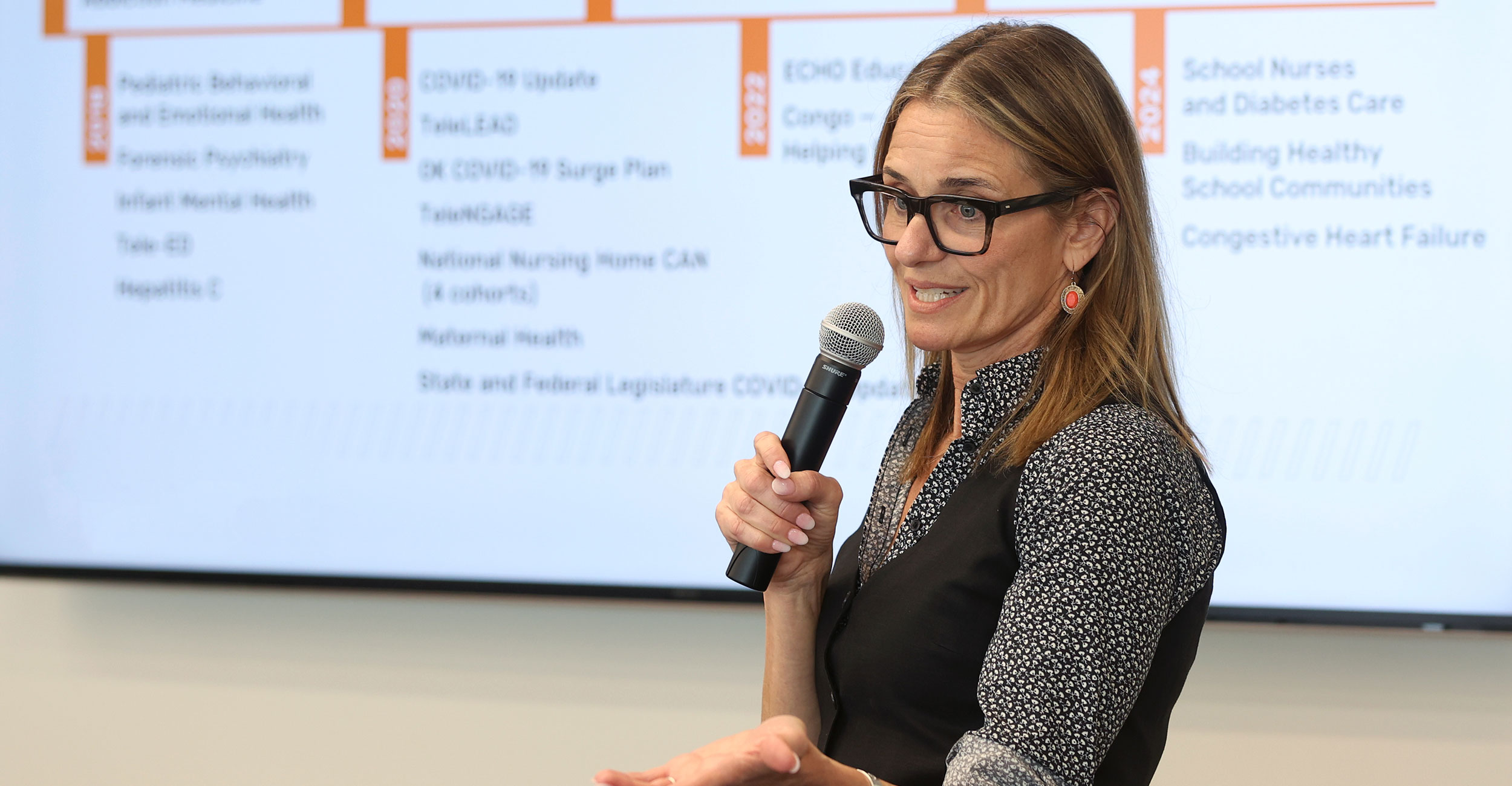 A speaker holds a handheld microphone while presenting in front of a projected slide listing health‑related topics, illustrating an educational or professional presentation setting.