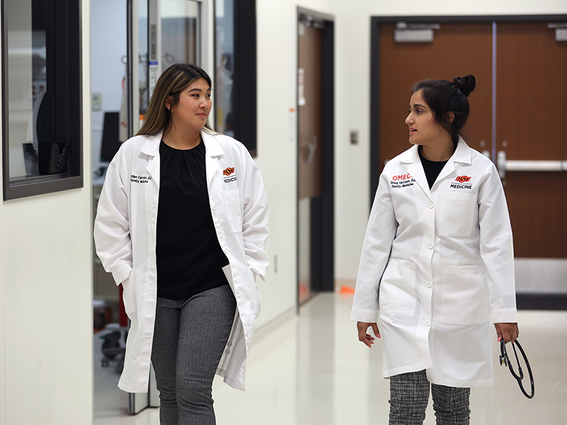 Two people wearing white medical lab coats walk together down a hallway in a clinical or educational facility, engaged in conversation.