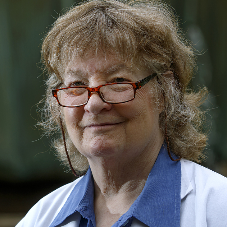 A professional headshot shows an individual wearing a white lab coat over a blue collared shirt, photographed outdoors with a softly blurred green background, conveying a healthcare or academic setting.