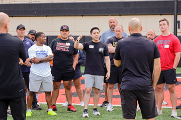 A coach addresses a group of participants standing on a football field, gesturing while leading an outdoor training or coaching session.